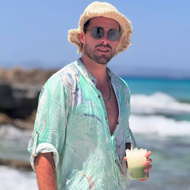 Hombre con camisa de acuarela en tonos verdes, sombrero y gafas de sol, disfrutando en la playa.
