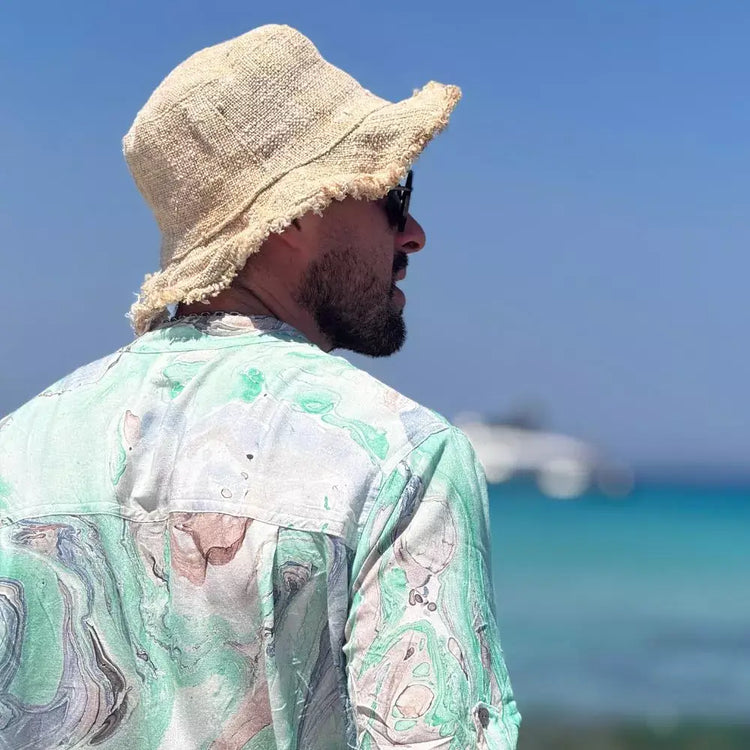 Hombre de espaldas usando una camisa de estampado acuarela y sombrero en una playa con mar azul de fondo.
