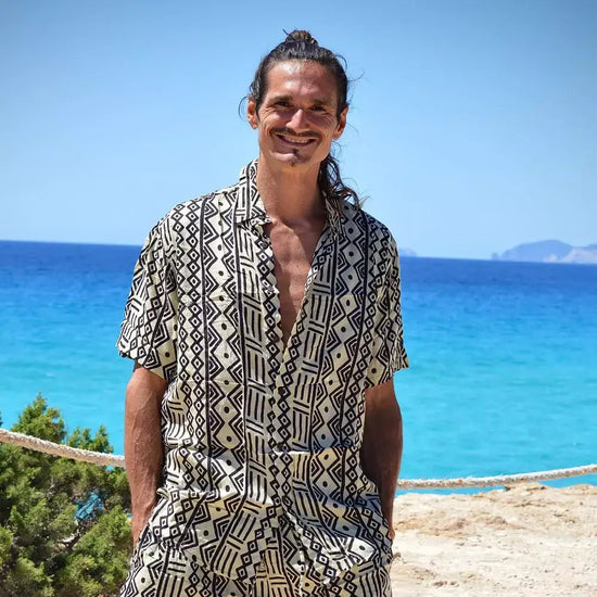 Hombre sonriente usando una camisa de rayón negra con estampado en una playa frente al mar azul.