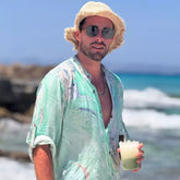 Hombre con camisa de acuarela en tonos verdes, sombrero y gafas de sol, disfrutando en la playa.