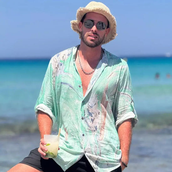 Hombre con camisa estampada en tonos verdes y sombrero, disfrutando de un día en la playa.
