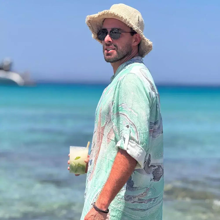 Hombre luciendo una camisa de rayon verde estilo acuarela junto al mar, disfrutando de una bebida refrescante.