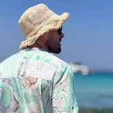 Hombre de espaldas usando una camisa de estampado acuarela y sombrero en una playa con mar azul de fondo.